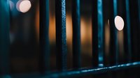 Dark moody composition with metal fence bars in foreground and soft glowing bokeh lights creating atmospheric depth and contrast between sharp and blurred elements.