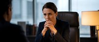 Professional businesswoman in dark suit sitting at office desk during serious meeting or interview with focused expression and clasped hands in modern corporate environment.
