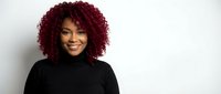 Young African American woman with curly burgundy hair smiling confidently in black turtleneck sweater against white background for professional headshot.