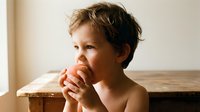 Young boy with light brown hair and fair complexion eating a red apple, seated at wooden table with neutral background, suitable for nutrition, health, children's education, and wellness content.