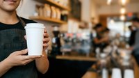Close-up of male barista in dark apron holding blank white takeaway cup in busy cafe environment with blurred background and warm lighting.