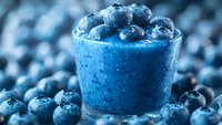 Fresh blueberry smoothie in glass with ripe berries on top surrounded by scattered blueberries for healthy breakfast drink and nutrition concept.