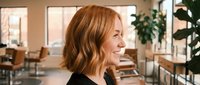 Professional woman with wavy red hair smiling in modern office workspace with natural lighting and plants creating warm productive atmosphere.
