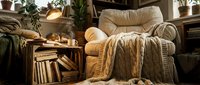 Cozy reading nook with upholstered armchair, quilted throw blanket, wooden side table stacked with books, potted plants, and warm lighting for relaxation and study.