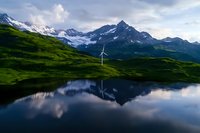 Wind turbine on green mountain slope reflected in alpine lake with snow-capped peaks and dramatic clouds in background, showcasing renewable energy in nature.