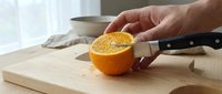 Male hand cutting fresh orange in half on wooden cutting board with knife and bowl in kitchen workspace.