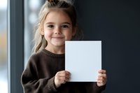 Young caucasian girl in brown sweater holding blank white paper card with gentle smile, looking at camera. Natural window lighting creates soft portrait mood.