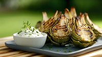 Grilled artichoke halves with creamy herb dip served on gray plate. Fresh Mediterranean appetizer garnished with chopped herbs and seasonings.