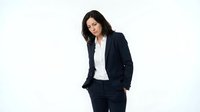 Professional woman with dark hair and olive complexion wearing formal black blazer and white shirt, standing against white background for corporate and business use.