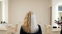 Woman with half blonde curly and half straight platinum hair sitting in modern salon chair showing hair transformation before and after styling treatment.