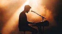 Silhouette of male musician performing at piano with microphone against dramatic orange stage lighting and atmospheric smoke effects creates moody concert atmosphere.