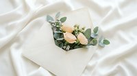 Elegant cream envelope with peach roses and eucalyptus branches on soft white fabric background for wedding invitations and romantic stationery designs.