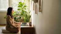 Young Asian woman in beige sweater sitting by window with green houseplants, reading book in cozy home interior with natural light and peaceful atmosphere.