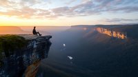 Silhouette of person sitting on cliff edge overlooking majestic mountain valley at sunset, dramatic landscape with golden light illuminating rocky cliffs and misty canyon below.