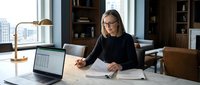 Professional businesswoman working at modern office desk reviewing documents with laptop computer in contemporary workspace with city view.