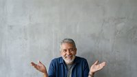 Mature man with gray hair and beard, wearing navy blue shirt, gesturing with open palms in shrug pose against textured concrete wall background for business and lifestyle content.