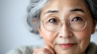 Thoughtful senior Asian woman with gray hair and round glasses, close-up portrait on light background, calm and wise expression.