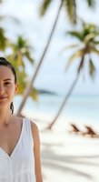 Young woman with dark hair and fair complexion wearing white sleeveless top on tropical beach with palm trees and ocean view for travel and lifestyle content.