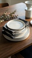 Stacked white ceramic plates with black napkins on wooden dining table, styled with eucalyptus and ceramic cups for table setting and home decor.