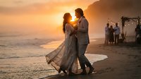 Romantic couple dancing on beach at sunset during wedding celebration with guests and ocean waves in background creating intimate moment for love story.