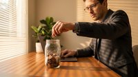 Young professional man in glasses saving coins in glass jar at wooden table in modern office interior with natural sunlight and plants.