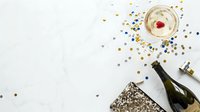 Champagne glass with gold and silver confetti scattered on white surface, black bottle and sequined clutch nearby, celebration party setup.