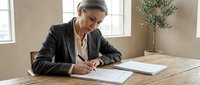 Mature businesswoman with gray hair signing documents at wooden desk in bright office space with natural lighting for corporate and professional use.