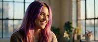 Young Caucasian woman with vibrant pink purple hair smiling in sunlit modern interior, warm natural light, casual knit sweater, positive lifestyle mood.
