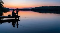Silhouette of male angler fishing from wooden dock at sunrise on calm lake, surrounded by forested shoreline, peaceful morning scene for outdoor recreation and leisure activities.