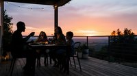 Silhouettes of diverse family gathered around outdoor dining table on wooden deck at sunset with scenic valley view and string lights overhead.