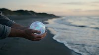 Person holding crystal ball on beach at sunset with ocean waves and coastal landscape reflecting magical light for meditation and spiritual concepts.