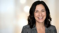 Professional middle-aged businesswoman with dark wavy hair smiling confidently in gray suit jacket against soft blurred office background for corporate use.