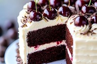 Chocolate layer cake with vanilla buttercream frosting and fresh cherries on top, cut to show moist interior and cream filling. Dark chocolate sponge layers.