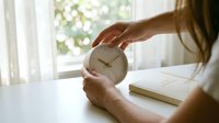 Woman holding analog clock at desk by window with notebook, representing time management, productivity, and scheduling concepts for business and lifestyle use.