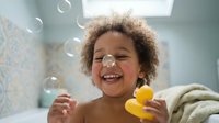 Joyful mixed-race toddler laughing in bathtub surrounded by soap bubbles, holding rubber duck, candid childhood bath time fun.