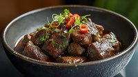 Tender braised beef chunks in rich brown sauce garnished with fresh microgreens and chili in black ceramic bowl, dramatic lighting creates appetizing mood.