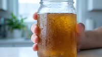 Unrecognizable person holding glass jar filled with cold amber beverage, condensation droplets on exterior, bright kitchen setting with blurred background and potted plant.
