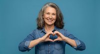 Smiling mature woman with gray hair making heart shape with hands wearing denim shirt on blue background expressing love and positivity.
