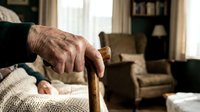 Elderly person's wrinkled hands gripping wooden walking cane in cozy living room interior. Senior care, aging, independence, mobility assistance concept.