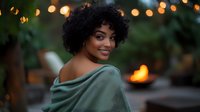 Young African American woman with curly hair smiling over shoulder in cozy green sweater with warm bokeh lights creating romantic evening atmosphere.
