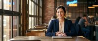 Professional businesswoman in navy blazer sitting at wooden table in modern office cafe with brick walls and large windows during daytime meeting.