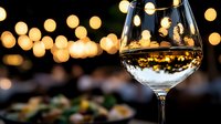 White wine glass reflecting golden bokeh lights against dark background with blurred food setting creates romantic dining atmosphere and celebratory mood.