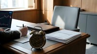 Professional businessman analyzing financial data and stock market charts on laptop in modern office workspace with documents and vintage clock.