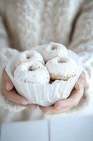 Powdered sugar donuts nestled in white paper liner held by feminine hands wearing cozy cream sweater, creating soft and inviting food photography composition.