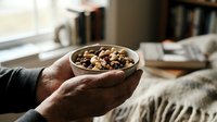 Hands holding wooden bowl filled with mixed nuts and dried fruits in cozy home setting with warm natural lighting for healthy snacking concept.