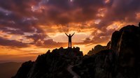 Silhouette of triumphant hiker with raised arms celebrating success on mountain peak at dramatic golden sunset with vibrant orange and purple clouds.