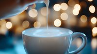 Pouring cream creates smooth swirl in coffee cup with creamy foam against blurred golden bokeh lights background, cozy morning atmosphere.