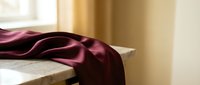 Luxurious burgundy silk fabric draped elegantly on white surface in warm sunlit interior, perfect for textile design and home decor projects.
