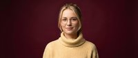 Young blonde Caucasian woman in yellow turtleneck sweater and glasses smiling confidently against burgundy background for professional headshot.