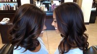 Dark wavy hair styled with soft curls and subtle brown highlights shown from two angles in salon setting, demonstrating professional hairstyling results.